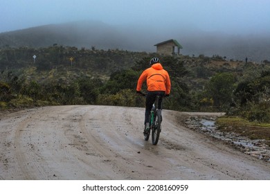 Cyclist Man With Orange Reflective Jacket Riding Mountain Bike On A Off Road Path In The Mountains. Background With A Cabin On The Fog And Moorland Vegetation