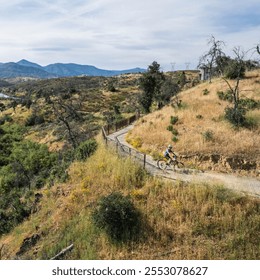 A cyclist maneuvers a winding trail through a dry, hilly landscape dotted with shrubs, under a wide sky with distant mountains and wind turbines in the backdrop - Powered by Shutterstock