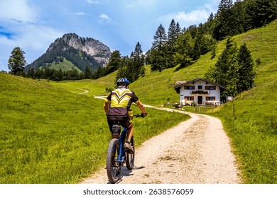 A cyclist navigates a winding dirt path surrounded by vibrant green fields and rolling hills, with a charming house in the background under a clear sky. - Powered by Shutterstock - Get 15% off with code: PIKWIZARD15