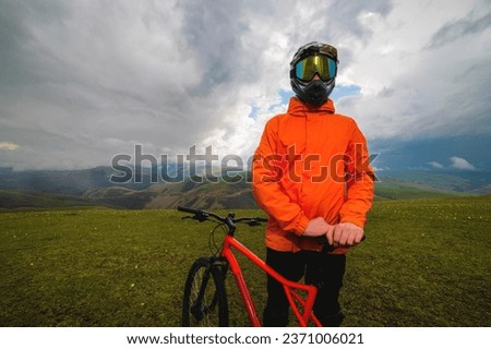 cyclist in an orange bright jacket and a protective helmet stands and holds a bicycle in the mountains, outside the city, off the road in summer in cloudy weather