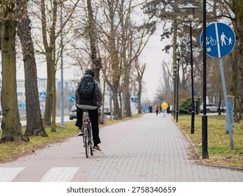 Cyclist rides down a paved path lined with trees. The path features a bicycle lane sign for pedestrians and cyclists. The cyclist wears a dark jacket and carries a backpack