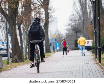 Cyclist rides down a paved path lined with trees. The path features a bicycle lane sign for pedestrians and cyclists. The cyclist wears a dark jacket and carries a backpack
