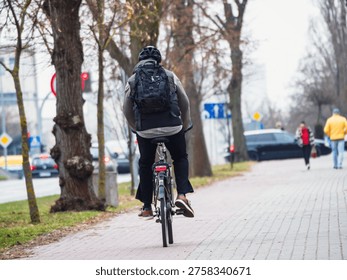 Cyclist rides down a paved path lined with trees. The path features a bicycle lane sign for pedestrians and cyclists. The cyclist wears a dark jacket and carries a backpack