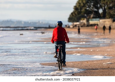 A Cyclist Riding Along Milford Beach In Auckland, With Out-of-focus People Walking And Dogs Playing On The Beach. 