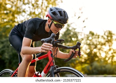 Cyclist Riding A Bike On An Open Road, Biking Cyclist Male Athlete Training Hard On Bicycle Outdoors At Sunset. Nature Landscape. Confident And Concentrated Man In Black Sportswear In Helmet