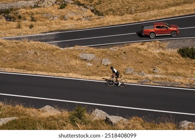 A cyclist riding uphill a winding road, with a pickup truck driving in the distance. The road is flanked by lush greenery, creating a scenic backdrop for the cyclist's journey. Romania - Powered by Shutterstock