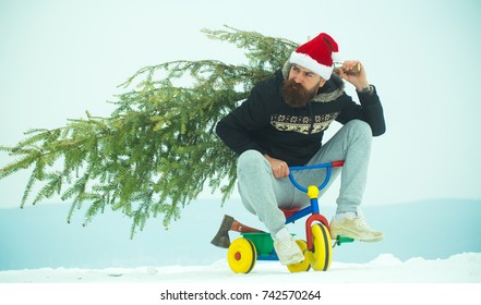 Cyclist In Santa Hat And Sporty Wear On Winter Day. Christmas And New Year. Hipster Carrying Xmas Tree And Axe On White Snow. Man Riding Tricycle On Snowy Landscape. Holiday Celebration Concept.