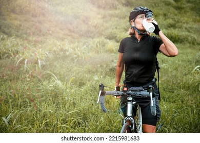 Cyclist senior woman drinking water from sports bottle in green grass meadow. Active Mature thirsty female in black sportswear outdoors. Copy space - Powered by Shutterstock