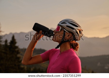Cyclist silhouette. Female cyclist in cycling clothes and a helmet drinks water from a sports bottle at sunset.
