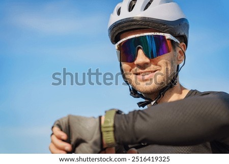 A cyclist smiles while checking his smartwatch, wearing a helmet and reflective sunglasses under a clear blue sky.
