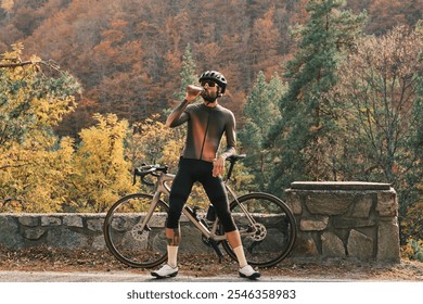 Cyclist takes a break in stunning autumn scenery, enjoying a refreshing drink. He stands with his road bike against a stone wall, surrounded by vibrant fall foliage. Romania. Carpathian Mountains. - Powered by Shutterstock