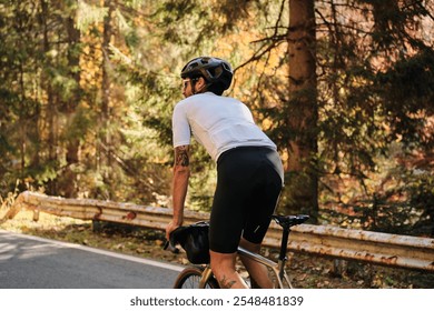 Cyclist in white jersey and black shorts rides through an autumnal forest. Sunlit road, scenic background. Perfect for active lifestyle, travel, and sports themes. - Powered by Shutterstock