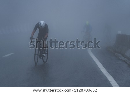 Cyclists are riding their bike up high on hill through a mist. Morning ride in a bad weather day: fog, rain, wind and cold day.