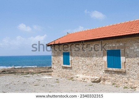 Cyprus stone house with blue shutters and red roof, blue sea in the background, pigion on the roof.