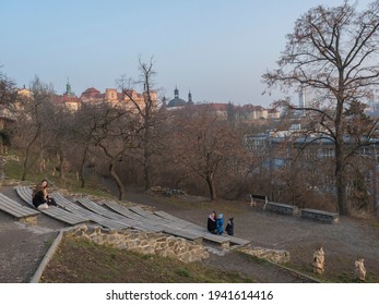 Czech Republic, Prague, February 23, 2021: Group Of People, Mother And Childern Sitting At Garden Albertovske Svahy With View Of Prague Karlov Quarter. Early Spring Day