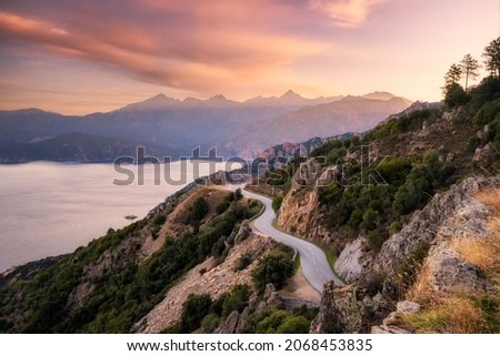 The D824 road winding its way along the coast from Capu Rossu towards Piana on the west coast of Corsica as the early  morning sun lights up the distant mountains