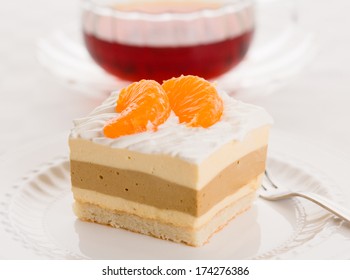 Dacquoise Cake Of Bavarian Cream, Coffee-flavored Hazelnut Praline, And Almond Meringue, Topped With Whipped Cream. Garnished With Pieces Of Mandarin Orange And Served With A Cup Of Tea.