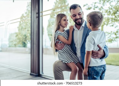 Dad Arrives Home From Work, His Little Son And Daughter Are Meeting Him. Happy Handsome Man Is Kneeling, Smiling And Hugging His Children.