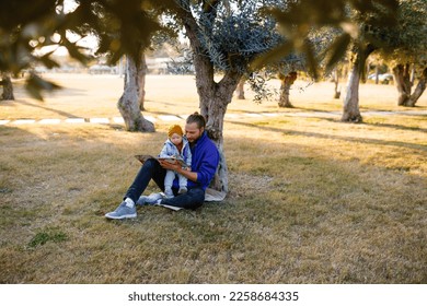 Dad and baby read a book under a tree in the park. Mediterranean landscape. - Shutterstock ID 2258684335