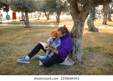 Dad and baby read a book under a tree in the park. Mediterranean landscape. - Shutterstock ID 2258684343