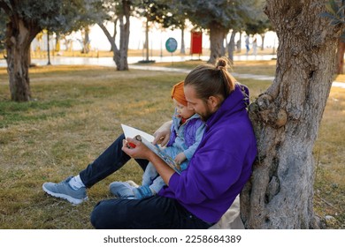 Dad and baby read a book under a tree in the park. Mediterranean landscape. - Shutterstock ID 2258684389