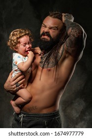 Dad Can't Handle A Crying Child, Guy Tears His Hair, The Baby's Tears Flow. Sporty Brutal Man With Beard And Tattoos Holds In His Arms A Little Curly-haired Boy.