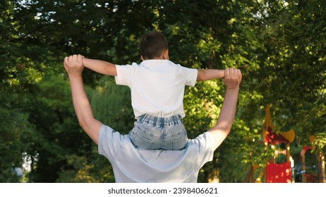 Dad carries infant boy on shoulders. Daddy wanders through city park with little son sitting atop shoulders. In expanse of trees in city park playful preschooler child sits in father shoulders