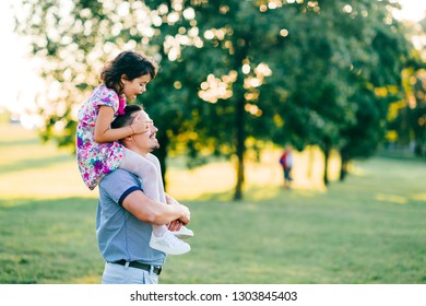 Dad carrying his daughter on shoulders in summer park at sunset. Father have fun with his little child. Kid siiting at parent neck. Happy family playing and making  crazy faces.  Lifestely portrait.