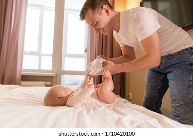 Dad Changes Baby Diapers Standing At Home In The Bedroom In Front Of The Child On The Bed