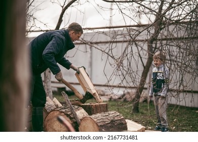 Dad Is Chopping Firewood In The Yard Of A Village House, The Boy Helps Dad Carry Firewood In The Woodpile. Dad And Son Work Together