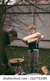 Dad Is Chopping Firewood In The Yard Of A Village House, The Boy Helps Dad Carry Firewood In The Woodpile. Dad And Son Work Together