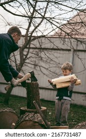 Dad Is Chopping Firewood In The Yard Of A Village House, The Boy Helps Dad Carry Firewood In The Woodpile. Dad And Son Work Together