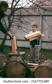 Dad Is Chopping Firewood In The Yard Of A Village House, The Boy Helps Dad Carry Firewood In The Woodpile. Dad And Son Work Together