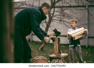 Dad Is Chopping Firewood In The Yard Of A Village House, The Boy Helps Dad Carry Firewood In The Woodpile. Dad And Son Work Together