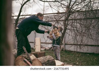 Dad Is Chopping Firewood In The Yard Of A Village House, The Boy Helps Dad Carry Firewood In The Woodpile. Dad And Son Work Together