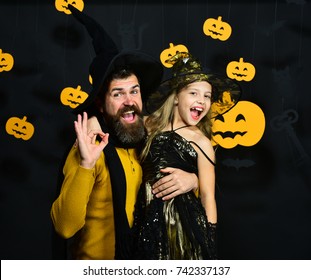 Dad And Daughter In Costumes. Halloween Party And Holiday Concept. Girl And Bearded Man With Cheerful Faces On Black Background With Decor. Wizard And Little Witch In Black Hats Hug And Show Ok Sign