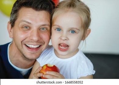 Dad And Daughter Laugh And Rejoice. Girl Blue-eyed Blonde With A Pigtail And A Red Apple. Family Happiness, Happy Smile, Joy Of Fatherhood.