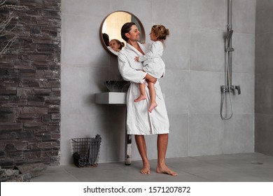 Dad And Daughter Looking At Each Other And Smiling. Family In Bathroom In Morning. Handsome Happy Father In White Bathrobe Holding Pretty Daughter In Pajama. Side View