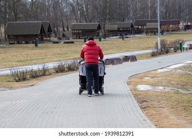 A Dad With A Double Pram Strolls Through A Cold, Deserted City Park In Early Spring. A Man In A Bright Red Warm Jacket.