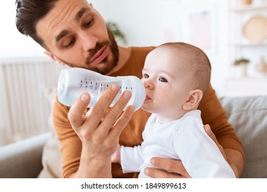 Dad Giving Bottle Of Water To Baby Toddler Feeding Him Sitting On Couch At Home. Kid's Hydration, Healthy Liquid For Your Child. Paternity Leave Fatherhood And Child Care, Parenthood. Selective Focus
