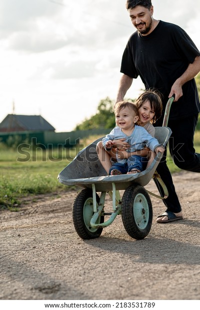 Dad has fun with the kids on\
the street on a summer day. The father drives the children in a\
wheelbarrow through the countryside. The concept of a happy\
childhood