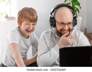 Dad In Headphones And Son Laughing Having Fun And Playing Video Game On The Computer At Home. Cheerful Family Spending Time Together Look At The Laptop Screen, Video Chatting With Mom, Friends