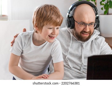 Dad In Headphones And Son Smiling Having Fun And Playing Video Game On The Computer At Home. Cheerful Family Spends Time Together, Communicates Via Video Chat With Mom, Friends