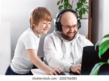 Dad In Headphones And Son Smiling Having Fun And Playing Video Game On The Computer At Home. Friendly Family Spends Time Together, Communicates Via Video Chat With Mom, Friends