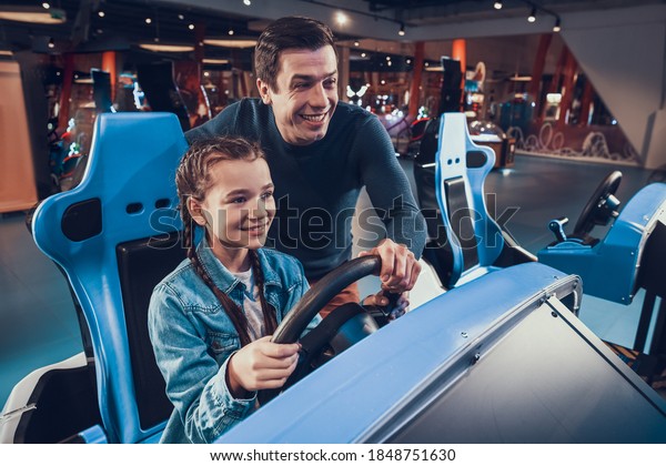 Dad helps\
his daughter drive a computer game. Little girl sits at the game\
wheel of the machine and drives the car.\
