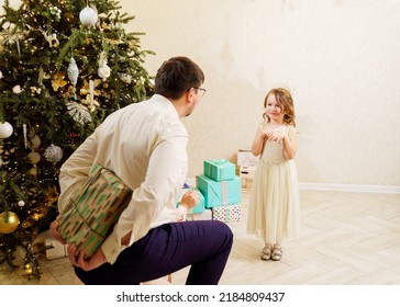 Dad Hides Behind His Back A Box With A Gift For The Baby Daughter. Father's Love And Care. The Tradition Of Giving Gifts For The New Year And Christmas.