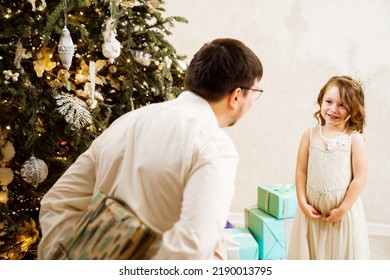 Dad Hides Behind His Back A Box With A Gift For The Baby Daughter. Father's Love And Care. The Tradition Of Giving Gifts For The New Year And Christmas.