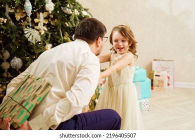 Dad Hides Behind His Back A Box With A Gift For The Baby Daughter. Father's Love And Care. The Tradition Of Giving Gifts For The New Year And Christmas.