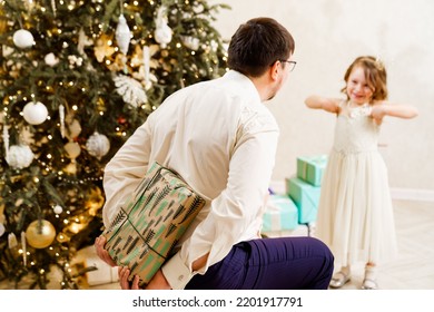 Dad Hides Behind His Back A Box With A Gift For The Baby Daughter. Father's Love And Care. The Tradition Of Giving Gifts For The New Year And Christmas.