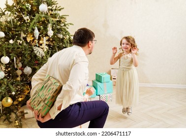 Dad Hides Behind His Back A Box With A Gift For The Baby Daughter. Father's Love And Care. The Tradition Of Giving Gifts For The New Year And Christmas.